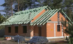 Budowa domu systemem gospodarczym w lesie. Dom z cegły, z zieloną więźbą dachową, w stanie surowym. Na placu budowy materiały budowlane.