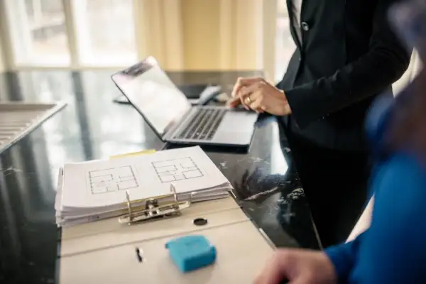 Plan mieszkania i laptop na blacie. Dwie osoby omawiają szczegóły, być może dotyczące ubezpieczenia mienia ruchomego.