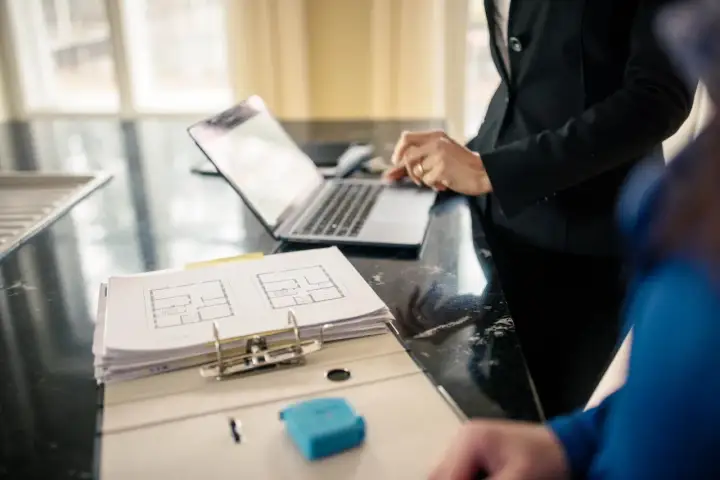 Plan mieszkania i laptop na blacie. Dwie osoby omawiają szczegóły, być może dotyczące ubezpieczenia mienia ruchomego.